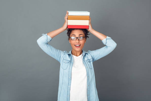 Young african female student isolated on grey wall wearing eyeglasses holding a pile of books on head looking camera fun